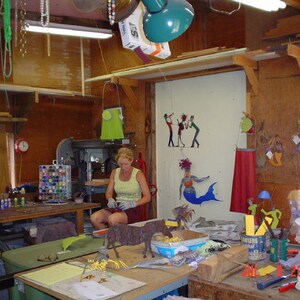 May include: A woman works in a workshop with a drill press and a table covered in tools and supplies. There are colourful metal sculptures on the wall, including a mermaid and a group of musicians. The year 2005 is written on the table.