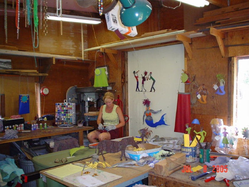May include: A woman works in a workshop with a drill press and a table covered in tools and supplies. There are colourful metal sculptures on the wall, including a mermaid and a group of musicians. The year 2005 is written on the table.