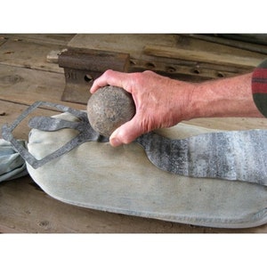 May include: A hand holds a round, dark grey stone over a metal cutout of a person. The cutout is on a light grey fabric surface.