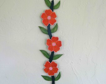 Könnte beinhalten: Vertikale florale Wandkunst mit drei orangefarbenen Blüten mit rosa Zentren und grünen Blättern. Die Blüten und Blätter sind an einem dunkelgrünen Stiel befestigt. Das Kunstwerk ist an einer strukturierten, cremefarbenen Wand befestigt.