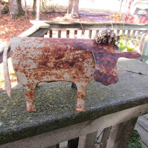 May include: A rusty metal cow sculpture with a white patina. The cow has a curly tail and a whimsical design.