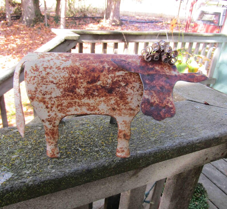 May include: A rusty metal cow sculpture with a white patina. The cow has a curly tail and a whimsical design.
