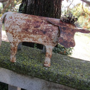 May include: A rusty metal cow sculpture with a curly tail and a white painted finish. The sculpture is sitting on a mossy surface.