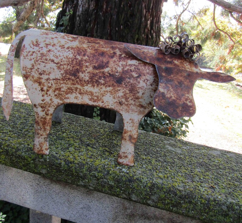 May include: A rusty metal cow sculpture with a curly tail and a white painted finish. The sculpture is sitting on a mossy surface.