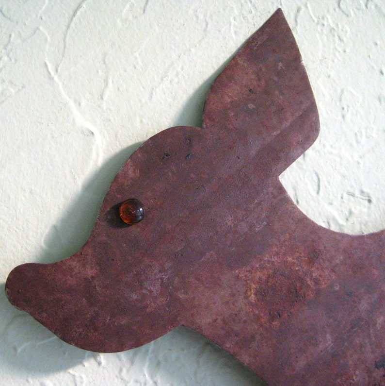 May include: A brown metal silhouette of a dog's head with a single amber glass eye. The metal has a rustic, aged look.
