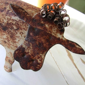 May include: A rusty metal sculpture of a cow with a curly tail made of wire. The cow is white with brown rust spots.