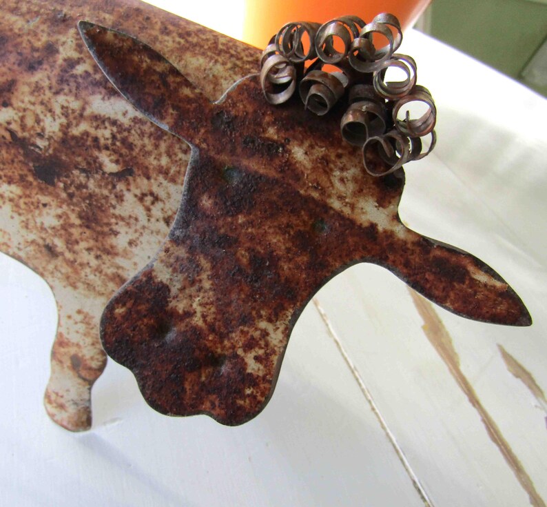 May include: A rusty metal sculpture of a cow with a curly tail made of wire. The cow is white with brown rust spots.