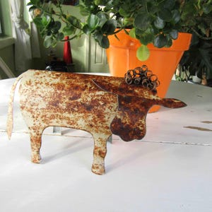 May include: A rusty metal cow sculpture with a white paint finish. The cow has a curled metal tail and horns. The sculpture is sitting on a white surface.