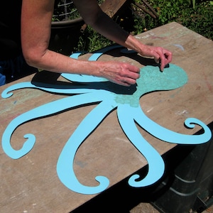 May include: A person is applying light blue paint to a large, blue, paper octopus cutout. The octopus has eight tentacles with curled ends. The octopus is on a wooden surface.