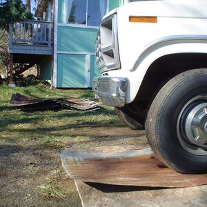 May include: A white pickup truck parked in front of a light blue building with a wooden deck. The truck has a chrome bumper and a large tyre. The truck is parked on a grassy area with a piece of metal on the ground.