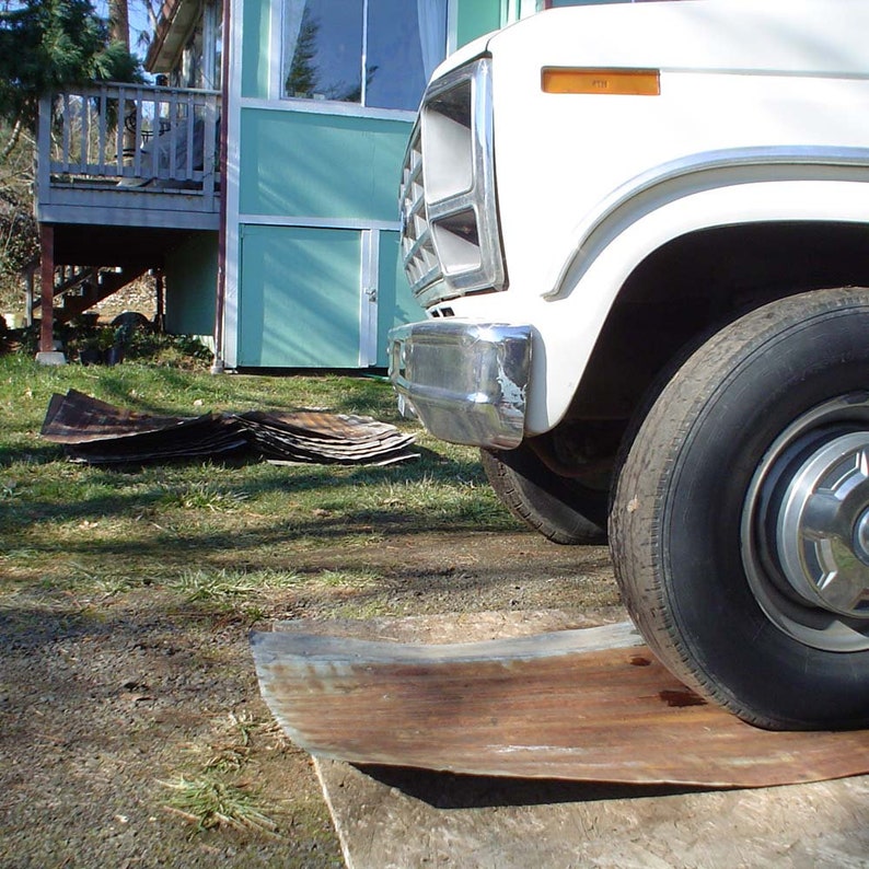 May include: A white pickup truck parked in front of a light blue building with a wooden deck. The truck has a chrome bumper and a large tyre. The truck is parked on a grassy area with a piece of metal on the ground.