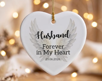 May include: A white heart-shaped ceramic ornament hanging from a gold string. The ornament features a graphic of two white angel wings and the black text: Husband, Forever in My Heart, and the date 25.06.2026 printed in the center.