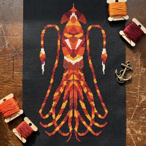 May include: A cross-stitch project on black fabric featuring a geometric squid design in shades of orange, red, and yellow. The piece is surrounded by four wooden spools of embroidery thread in matching colors, a single sewing needle, and a metal anchor.