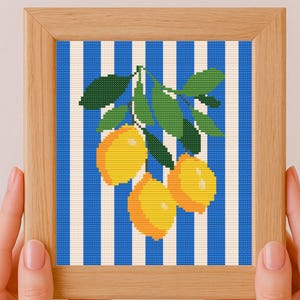 May include: A framed cross-stitch artwork featuring three yellow lemons with green leaves against a blue and white striped background. The artwork is in a light brown wooden frame.