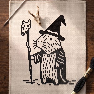 May include: A cross-stitch project on beige Aida fabric featuring a black thread design of a robed figure wearing a pointed hat and holding a staff. A silver needle, a small animal skull, and a skein of black embroidery floss are placed nearby.
