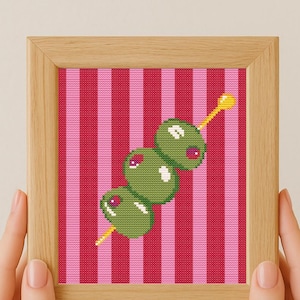 Puede incluir: Una obra de arte en punto de cruz enmarcada con tres aceitunas verdes con centros rojos en un palillo amarillo. El fondo tiene rayas verticales rosas y rojas. El marco es de madera clara.