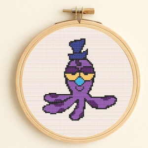 Può includere: Un disegno a punto croce di un polpo viola con un cappello a cilindro blu e occhiali da sole gialli, inserito in un telaio da ricamo in legno. Il polpo ha un naso blu e macchie nere sulle braccia.