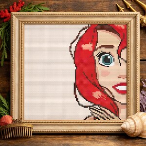 May include: A cross-stitch pattern framed in a light gold wooden frame, featuring the right half of a face with vibrant red hair, large blue eyes, and bright red lips, set against a plain white background within the stitched design area.