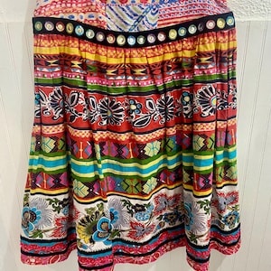 Puede incluir: Una falda colorida con múltiples patrones, una cintura negra y una fila de adornos de espejo. La falda tiene una variedad de diseños florales y geométricos en tonos rojos, amarillos, azules, verdes y rosas.