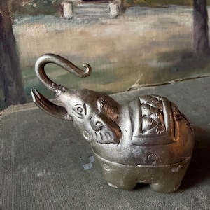 Op de afbeelding: Een zilverkleurig metalen olifantenbeeldje met een omhoog gekrulde slurf, puntige slagtanden en een decoratief gegraveerd zadelpatroon op de rug. Het object heeft een gladde, metalen afwerking met zichtbare gegraveerde details op de kop en het lichaam.