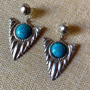 May include: A pair of silver earrings featuring a turquoise stone. The earrings have a leaf-shaped design with a textured finish and a post fastening. The turquoise stones are round and have a speckled appearance.