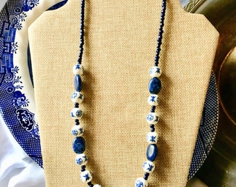 May include: A blue and white ceramic bead necklace with dark blue gemstone beads. The necklace is displayed on a brown fabric stand.