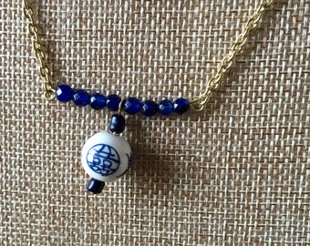 Könnte beinhalten: Eine goldene Kettenkette mit einem blauen und weißen Porzellananhänger. Der Anhänger zeigt ein chinesisches Schriftzeichen für doppeltes Glück. Die Kette ist mit dunkelblauen Perlen verziert.