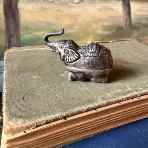May include: A small, metallic, silver-toned elephant figurine with an upward-curving trunk. The surface features intricate, embossed decorative patterns on the head, ears, and back, with a smooth, rounded body and visible legs in a seated position.