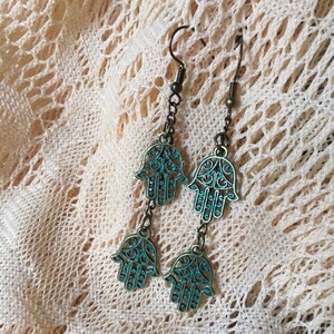 May include: A pair of dangle earrings with a turquoise blue hamsa hand charm. The earrings are made of metal and have a filigree design.