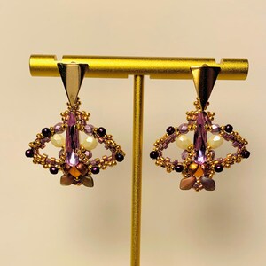 May include: A pair of dangle earrings featuring a gold-tone triangular top piece connected to a beaded moth-shaped design. The moth body is composed of a central lavender crystal, surrounded by small gold, purple, and cream-colored beads.