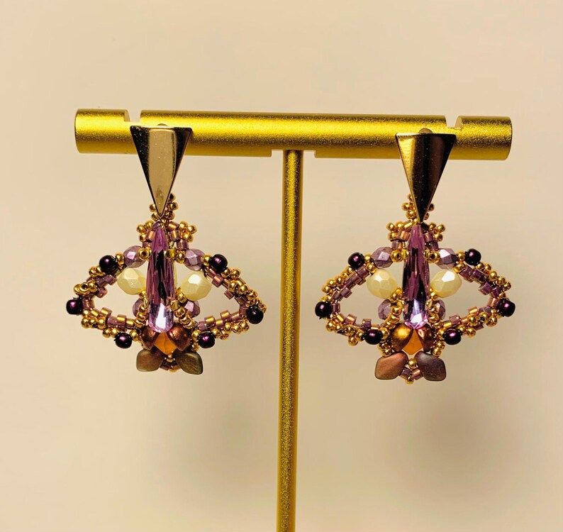 May include: A pair of dangle earrings featuring a gold-tone triangular top piece connected to a beaded moth-shaped design. The moth body is composed of a central lavender crystal, surrounded by small gold, purple, and cream-colored beads.