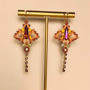 May include: A pair of dangle earrings featuring a central deep orange marquise crystal surrounded by small yellow, peach, and iridescent beads. Each earring has a dangling chain of small clear crystals and is suspended from a gold-tone hook.