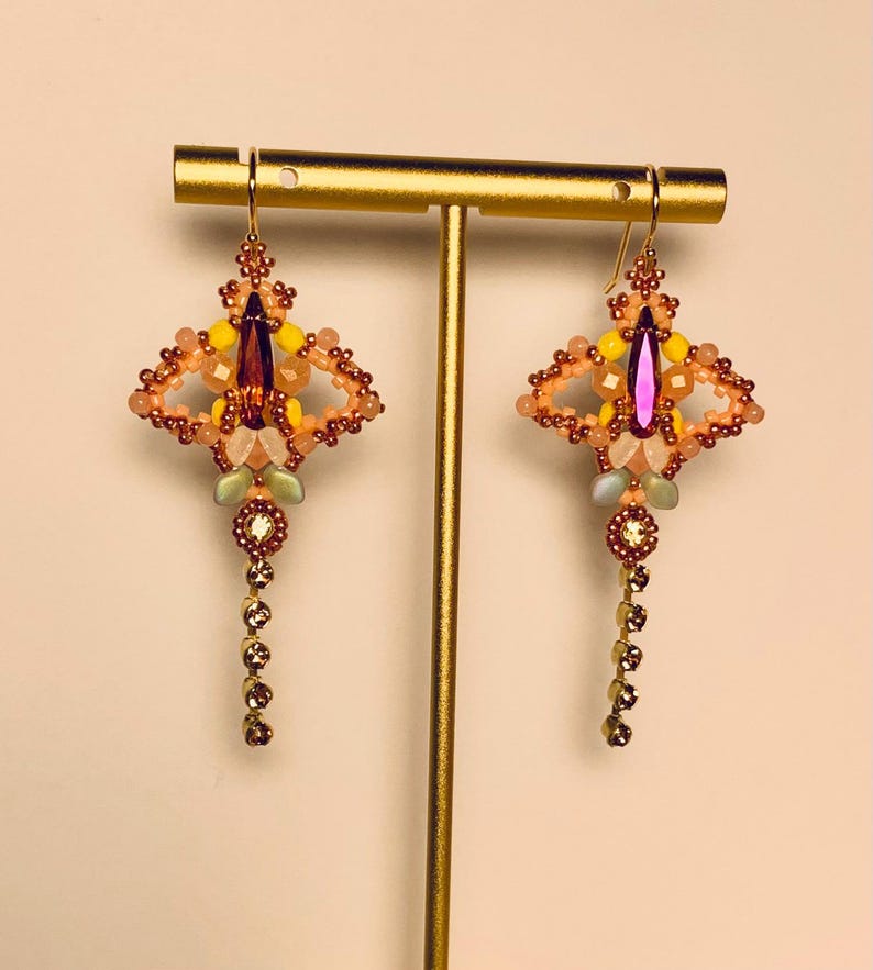 May include: A pair of dangle earrings featuring a central deep orange marquise crystal surrounded by small yellow, peach, and iridescent beads. Each earring has a dangling chain of small clear crystals and is suspended from a gold-tone hook.