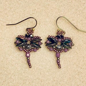 May include: A pair of dangle earrings with a geometric design. The earrings are made with purple, silver, and black beads. The earrings are attached to gold hooks.