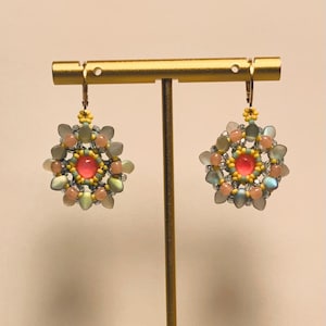 May include: A pair of circular beaded earrings featuring a central bright pink cabochon surrounded by concentric rings of small yellow, peach, and translucent blue-grey beads. The earrings are constructed with a floral-like pattern of petal-shaped beads.