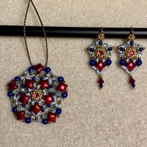 May include: A beaded jewellery set comprising a pendant and matching earrings. The pendant is a starburst design with red, blue, and gold beads. The earrings mirror the pendant's design, with a dangling red bead. The jewellery is displayed against a neutral background.