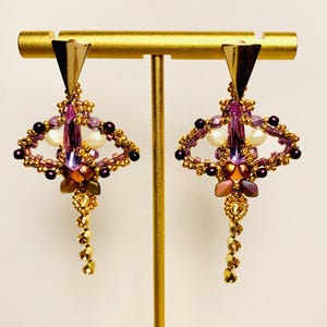 May include: A pair of dangle earrings featuring a gold-tone triangular top piece, a central elongated purple crystal, and intricate beadwork in shades of purple, gold, and cream. The design includes a dangling beaded chain at the bottom.