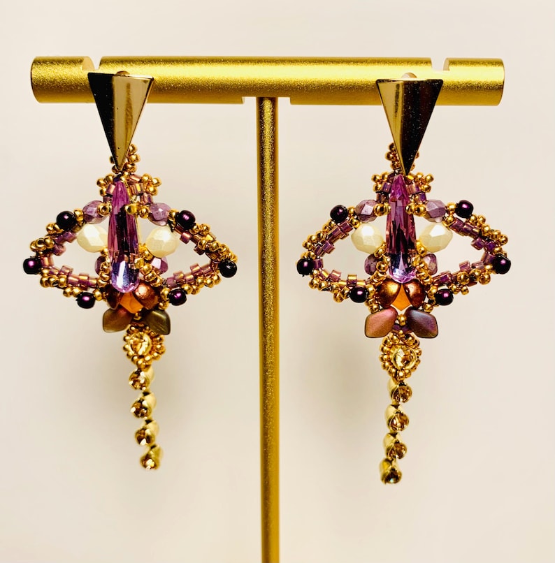 May include: A pair of dangle earrings featuring a gold-tone triangular top piece, a central elongated purple crystal, and intricate beadwork in shades of purple, gold, and cream. The design includes a dangling beaded chain at the bottom.