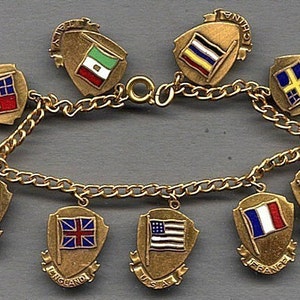 May include: A gold-tone charm bracelet featuring nine shield-shaped pendants, each displaying a colorful enamel flag and the name of a country including Canada, Italy, China, Sweden, Russia, England, USA, France, and Greece, connected by a link chain.