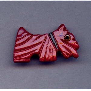 May include: A red, stylized Scotty dog brooch with a black collar and a gold-colored eye. The brooch has a glossy finish and is carved with textured lines. The dog is positioned on a gray background.