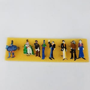 May include: Set of eight enamel pins featuring colorful figures in various poses. The pins are displayed on a yellow foam background.