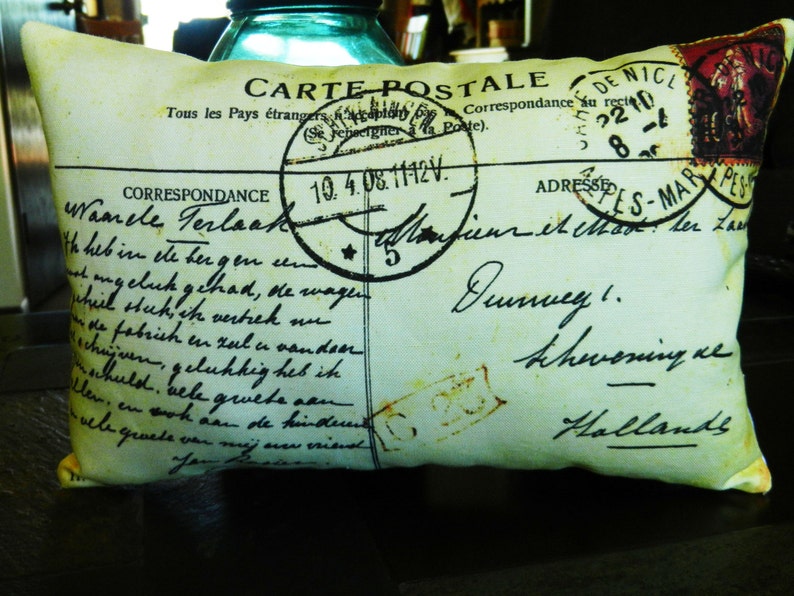 Puede incluir: Una almohada decorativa con un dise&ntilde;o de postal vintage. La almohada presenta un fondo verde descolorido con texto negro y un sello rojo, blanco y azul. El texto en la almohada dice "Carte Postale" y "Correspondance."
