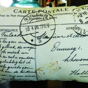 Puede incluir: Una almohada con forma de postal vintage con fondo crema y texto negro. El texto incluye las palabras "Carte Postale" y "Correspondance". La almohada tiene un aspecto desgastado y presenta un sello descolorido con el texto "De Nicl" y "2210 8-1".