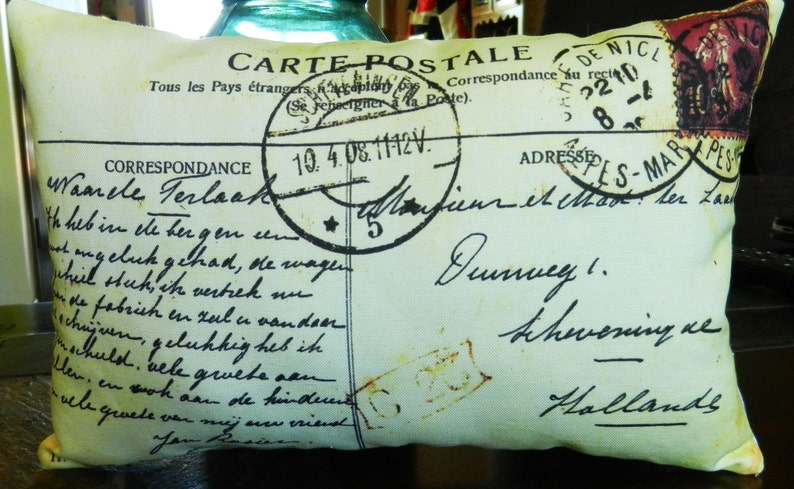Puede incluir: Una almohada con forma de postal vintage con fondo crema y texto negro. El texto incluye las palabras "Carte Postale" y "Correspondance". La almohada tiene un aspecto desgastado y presenta un sello descolorido con el texto "De Nicl" y "2210 8-1".