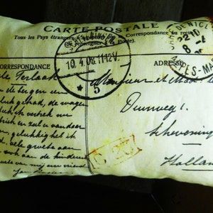 Puede incluir: Una almohada con forma de postal vintage con fondo amarillo y texto negro. El texto incluye las palabras "Carte Postale" y "Correspondance". La almohada tiene un aspecto desgastado y envejecido.