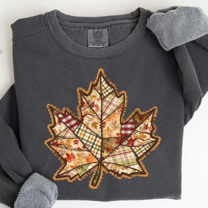 Peut inclure: Sweat-shirt gris foncé avec une grande feuille d'érable en patchwork aux couleurs automnales. La feuille présente divers motifs à carreaux et floraux. Le mot "PEPPER" est imprimé en bas.