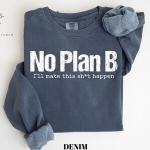 May include: A denim blue crewneck sweatshirt featuring the white distressed text No Plan B and the phrase I'll make this sh*t happen printed on the front. The garment has long sleeves with ribbed cuffs and a ribbed neckline, showcasing a casual style.