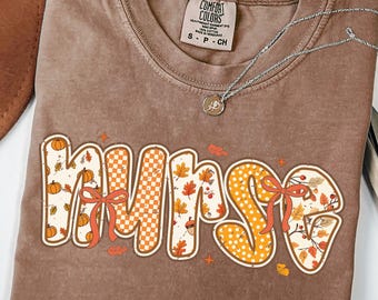 May include: Espresso-colored t-shirt with the word "NURSE" in a fall-themed design. The letters feature pumpkins, leaves, and bows in orange, white, and brown. A silver necklace rests on the shirt.