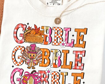 May include: Ivory-colored t-shirt with a Thanksgiving-themed graphic. The design features the words "GOBBLE GOBBLE GOBBLE" in orange, pink, and white checkered letters, along with illustrations of a pie, turkey, and pumpkin.