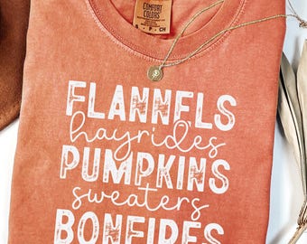 May include: Orange Comfort Colors t-shirt with the words "Flannels, hayrides, pumpkins, sweaters, bonfires" in white. A gold necklace and a brown hat are next to the shirt. The shirt has a tag that says "Comfort Colors".